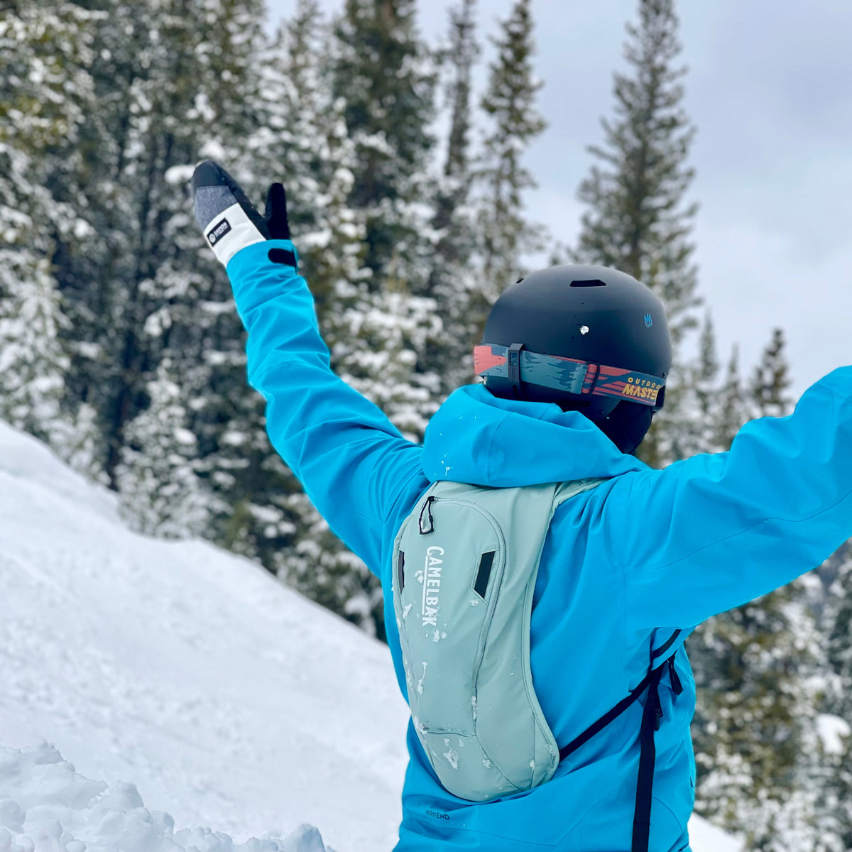 Skier in a blue jacket raising both arms on a snowy mountain slope with winter trees in the background, wearing helmet and gloves.