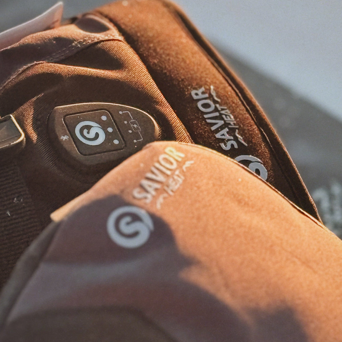 Close-up of a brown heated glove or mitten featuring the 'SAVIOR HEAT' logo and a control button with a spiral icon for adjusting warmth.