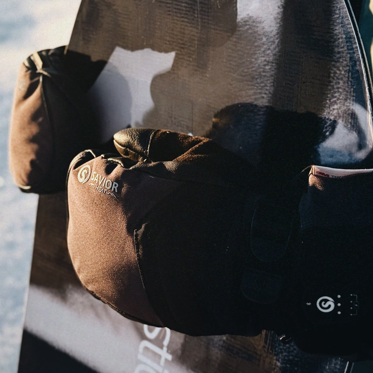 Brown heated mittens by SAVIOR HEAT, featuring a thumb compartment, integrated control button with spiral icon, and adjustable design for warmth during winter sports.