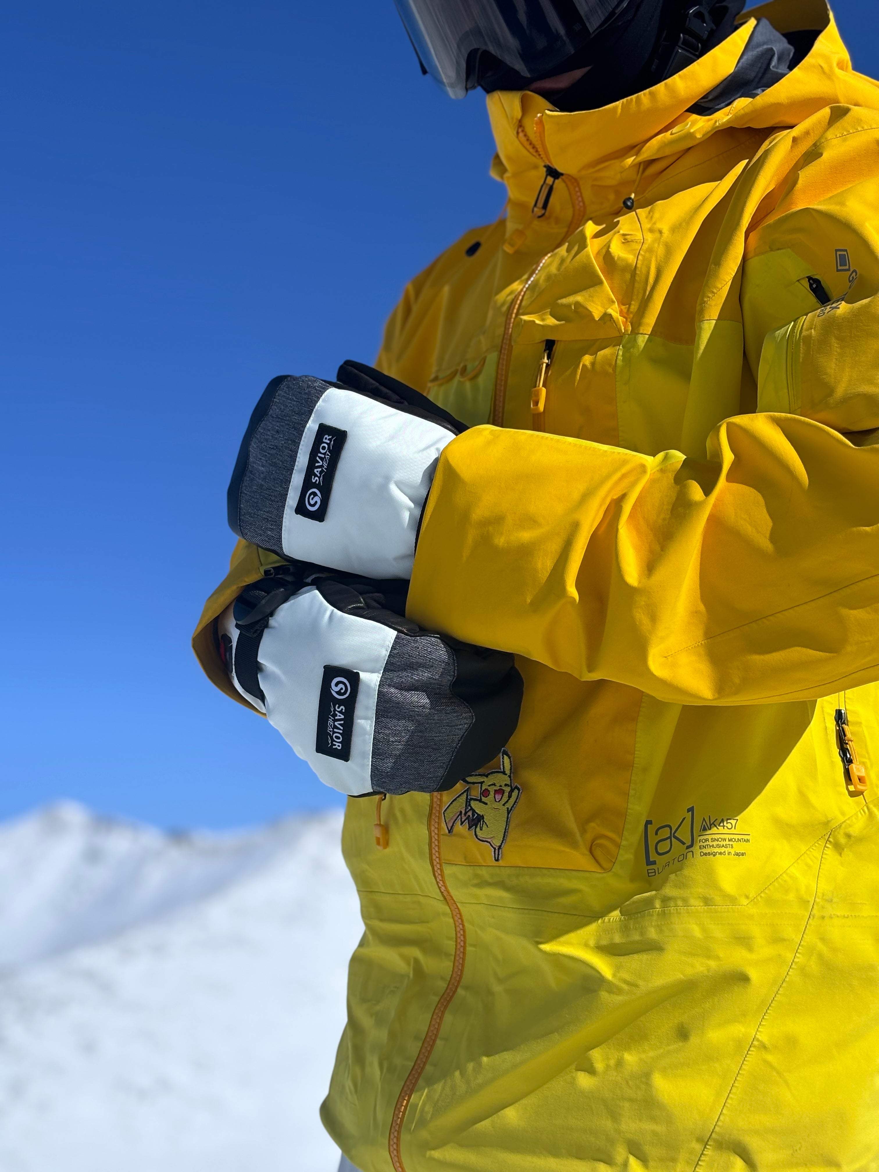 Skier in a yellow jacket adjusting battery heated gloves under bright alpine sun, highlighting winter warmth and high-performance outdoor gear.