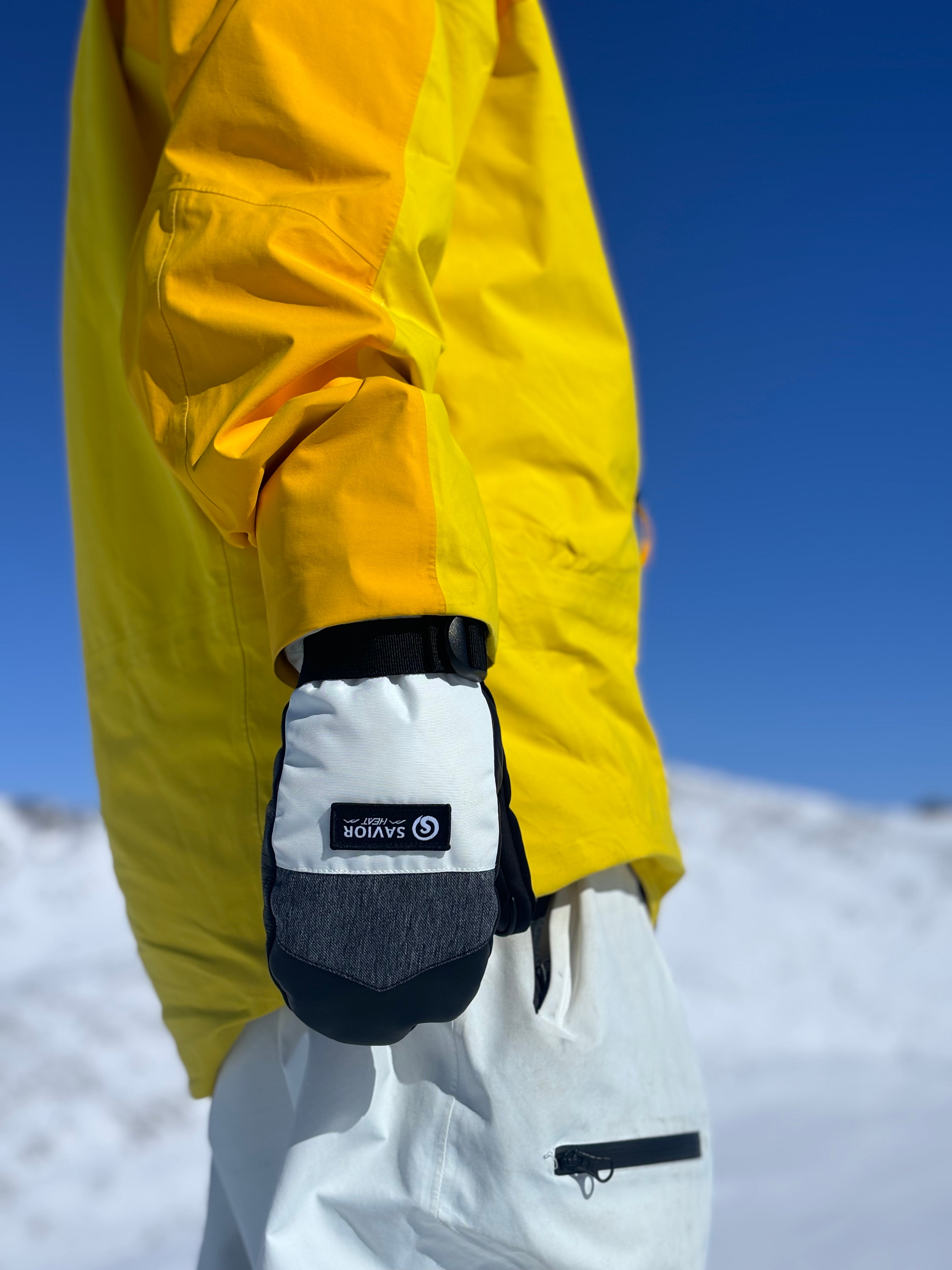 Snowboarder in a bright yellow jacket wearing battery heated gloves, standing in a sunny alpine landscape for winter warmth and comfort.