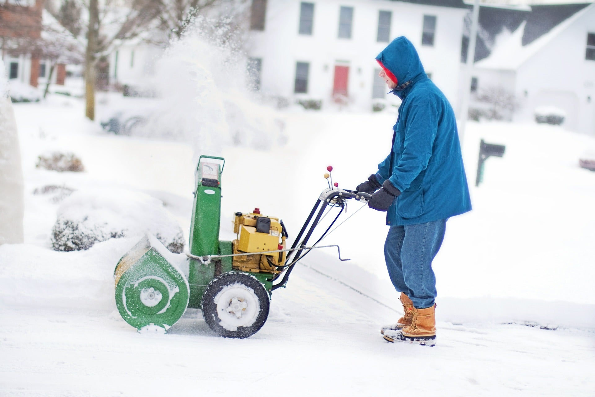 Man wearing insulated winter gloves operating a snow blower in heavy snow outside a residential home.