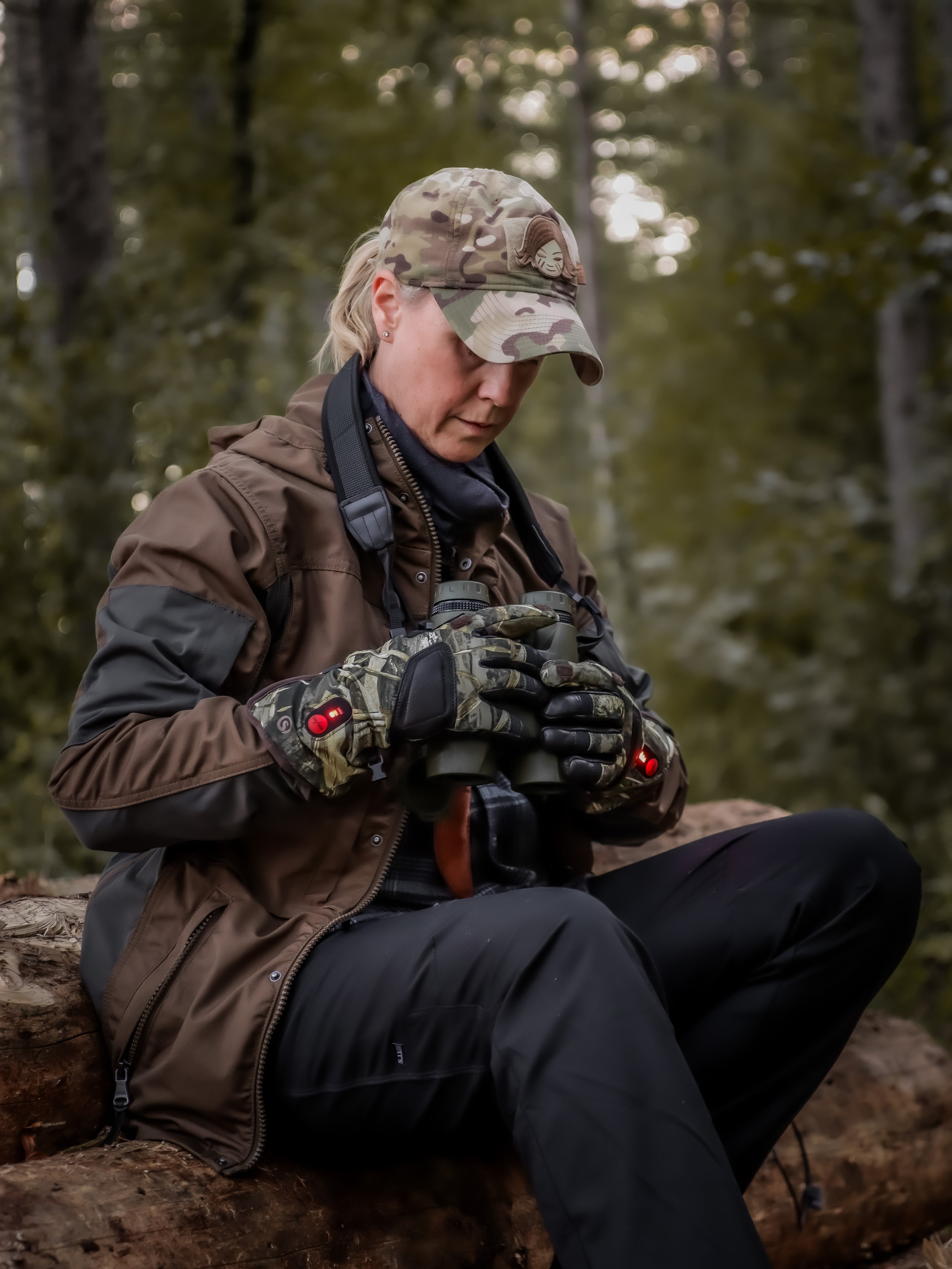 Woman hunter in forest wearing camo heated gloves while holding binoculars.