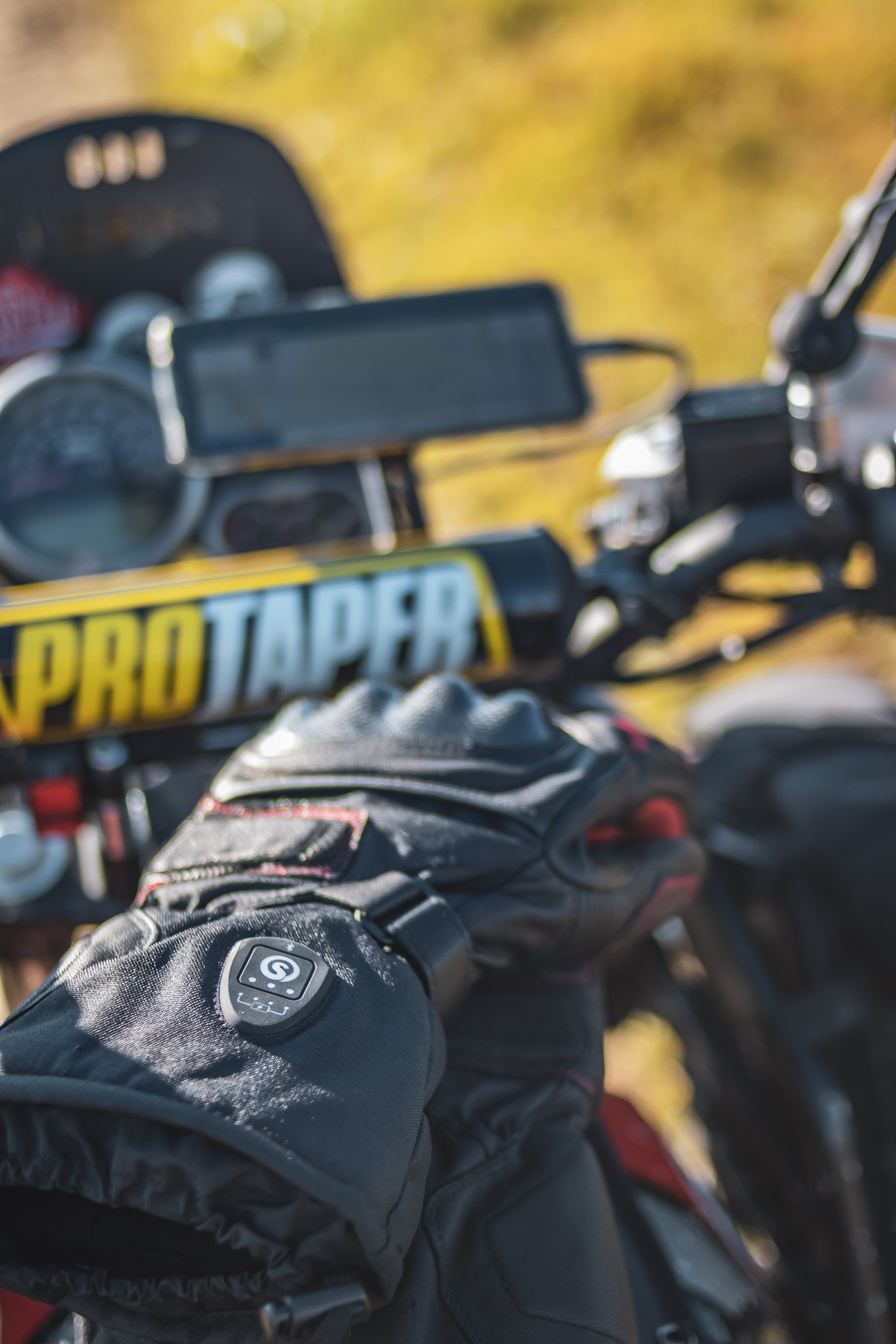 Heated motorcycle gloves in use during an outdoor ride, showing the control button and protective build on the handlebars.