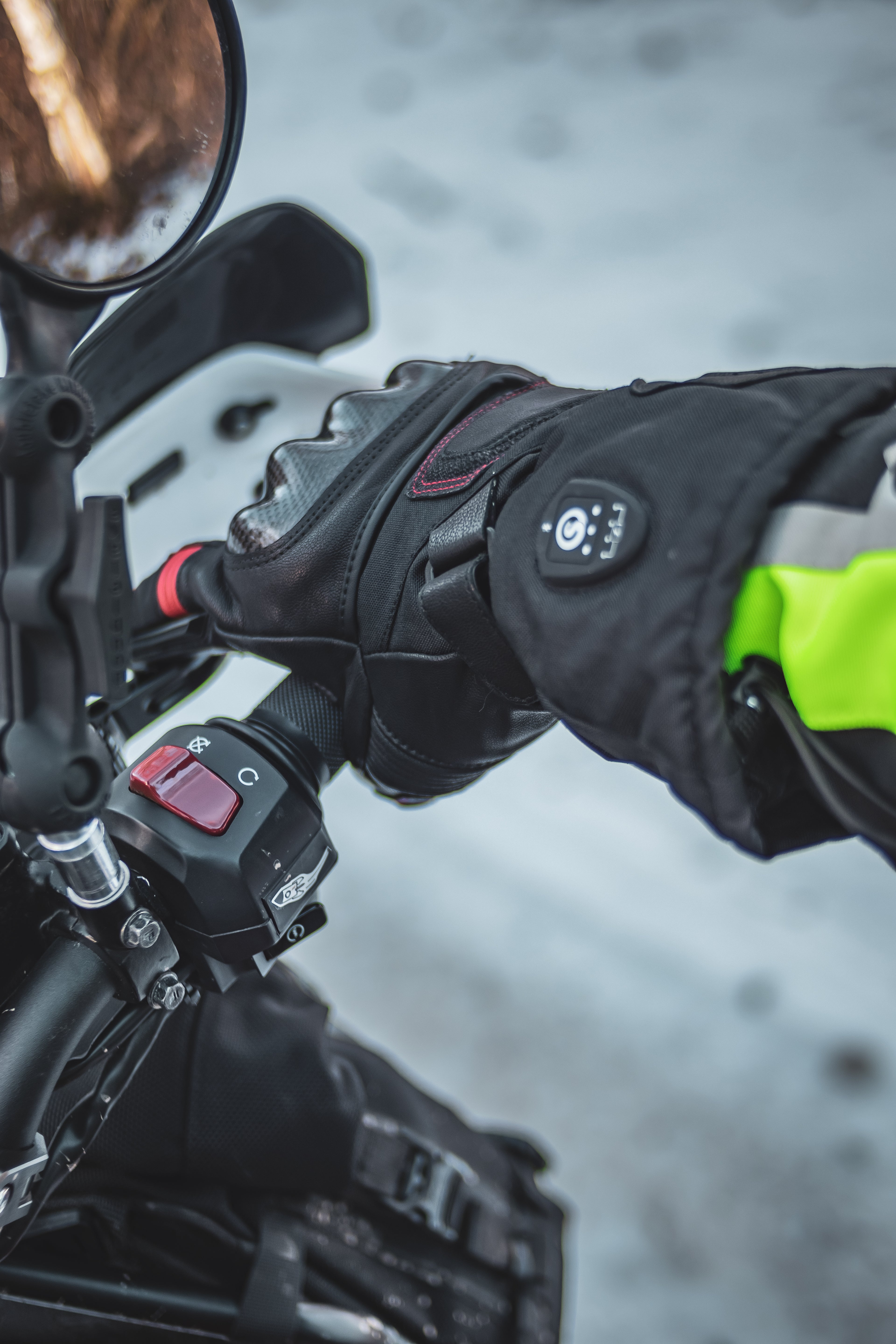 Rider gripping the motorcycle handlebar in winter conditions, wearing insulated heated gloves with a visible control button