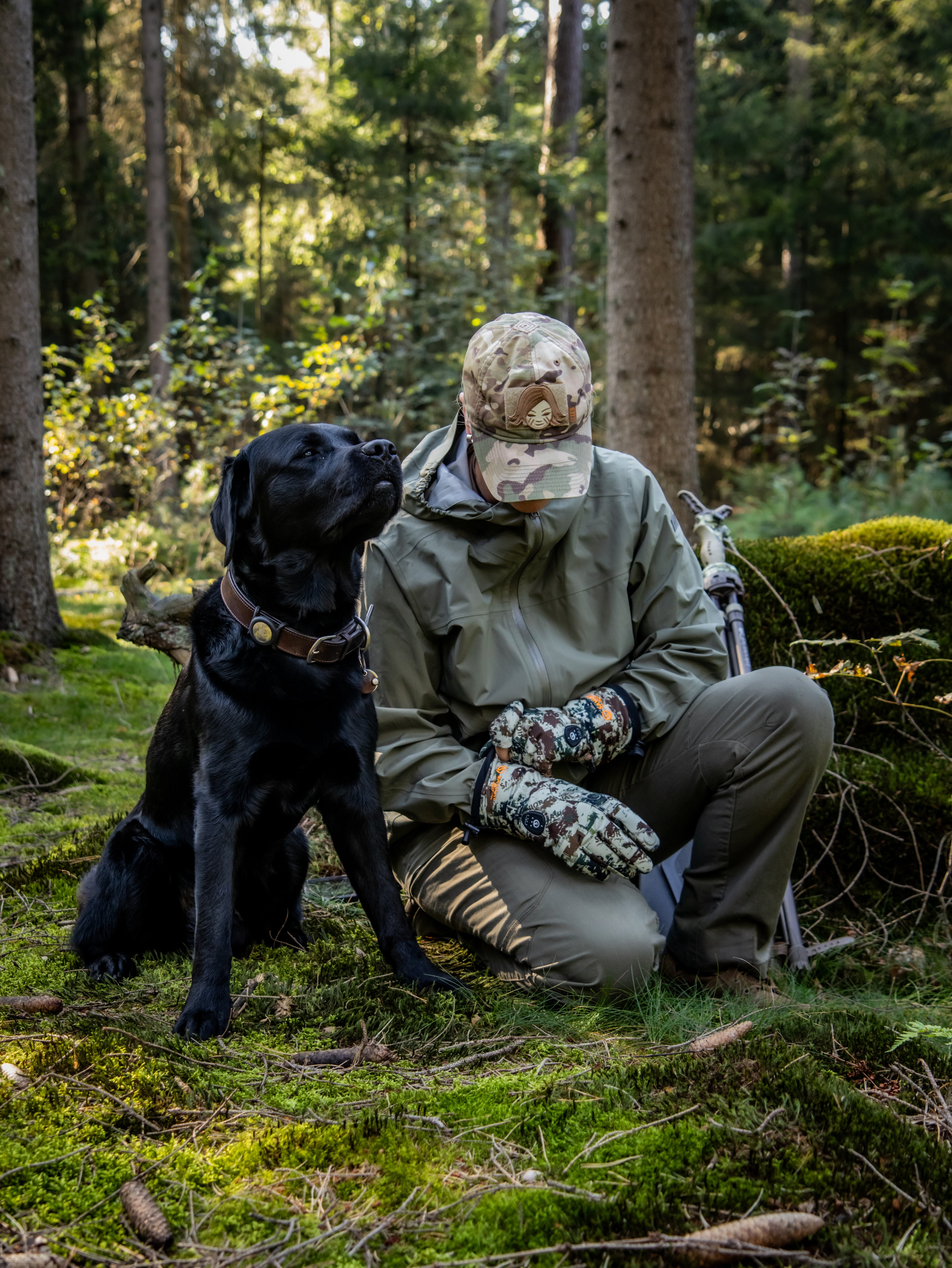 Hunter sitting in a forest wearing camo heated gloves beside a black Labrador.