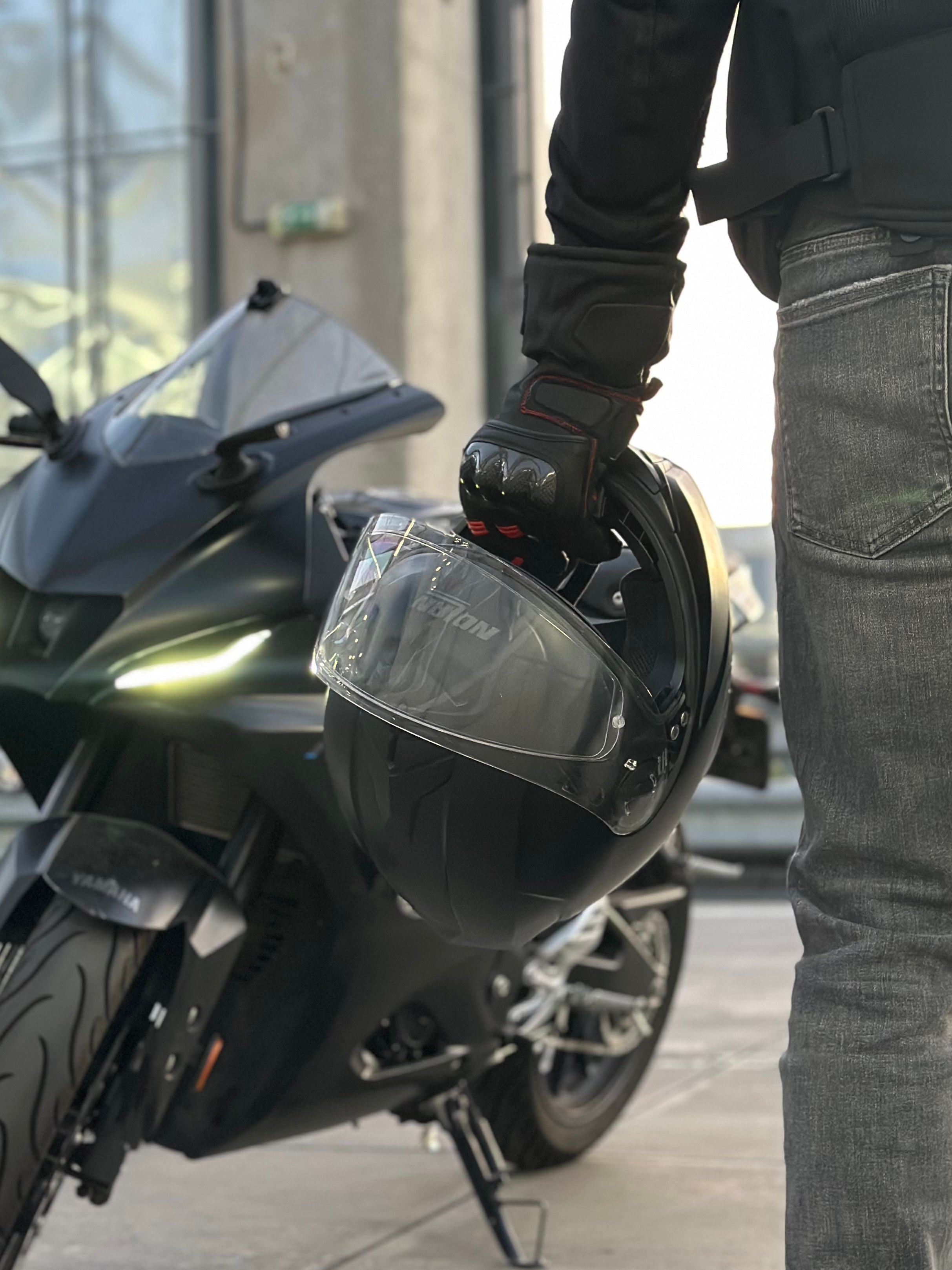 Motorcyclist standing beside a sport bike while holding a helmet, wearing protective riding gloves in an urban outdoor setting.
