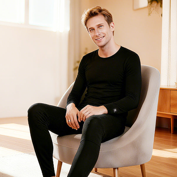 Man wearing black heated underwear set, seated indoors, showing slim fit long-sleeve top and pants designed for warmth.