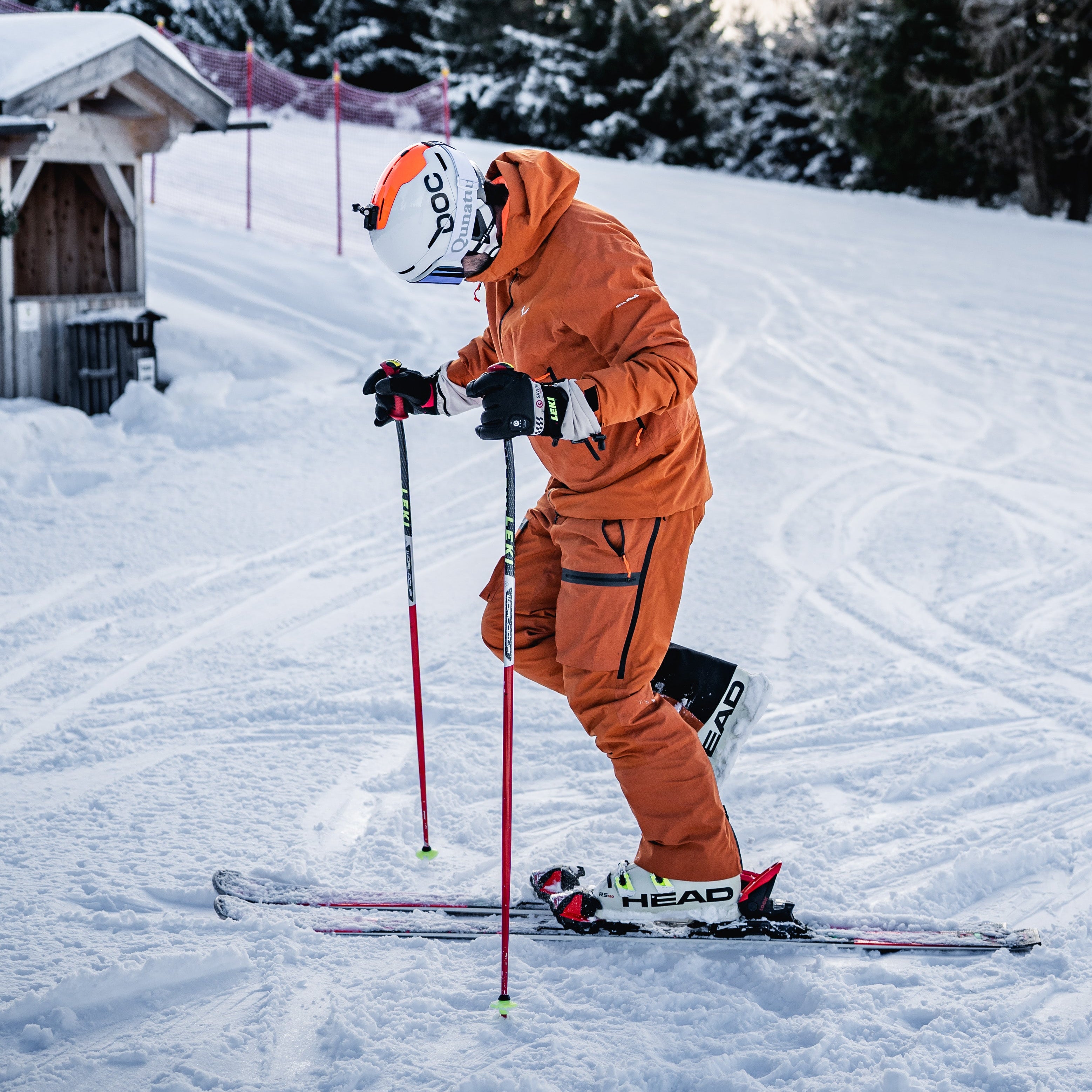 Skier in orange gear standing on snowy terrain, adjusting ski poles while wearing insulated gloves in a winter mountain setting.