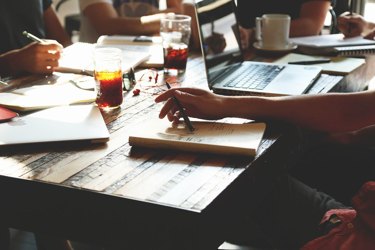 Startup team meeting at a wooden table with laptops, notebooks, and iced drinks as members brainstorm ideas and collaborate on project planning.